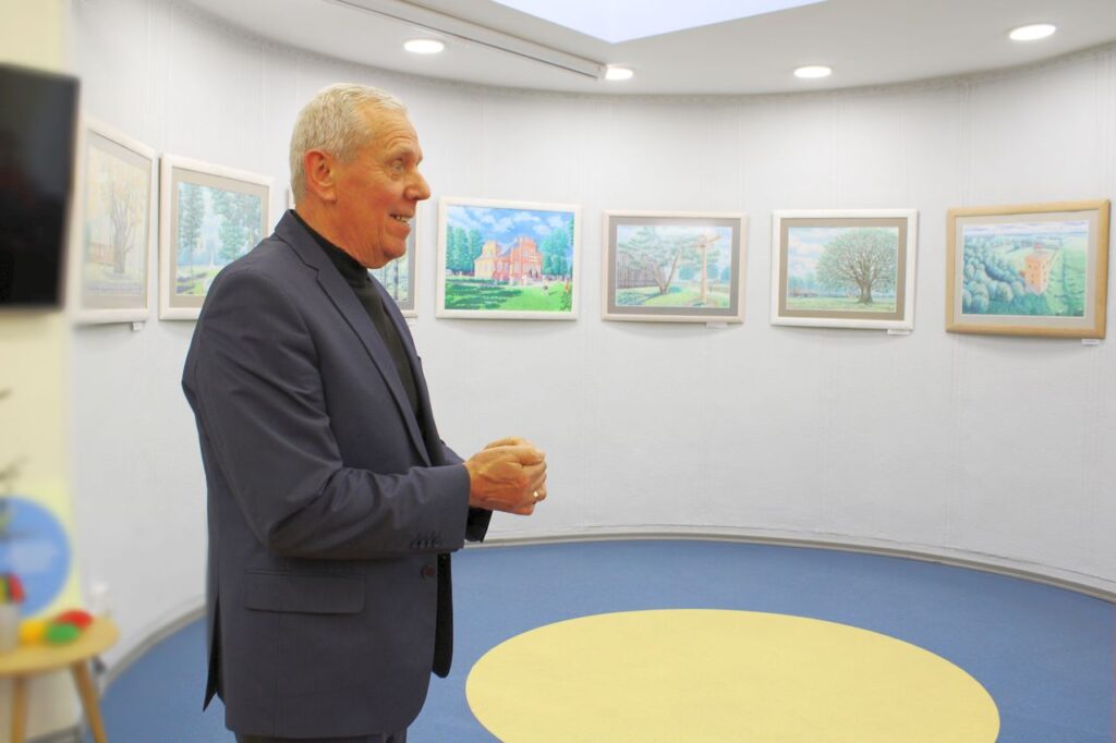 Tautodailininkas K. V. Henrikas Vaičiūnas bibliotekoje prie savo tapytų darbų