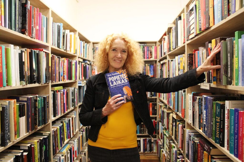 Moteris su knyga rankoje pozuoja prie knygų lentynų