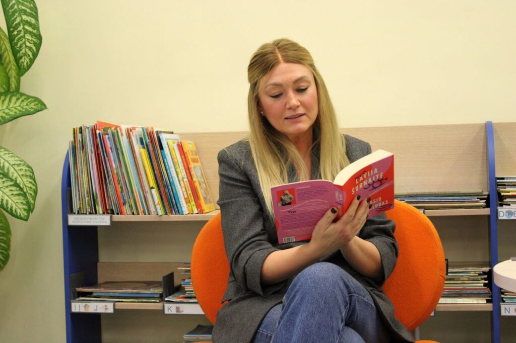 Moteris su atversta knyga rankose sėdi knygų lentynų fone