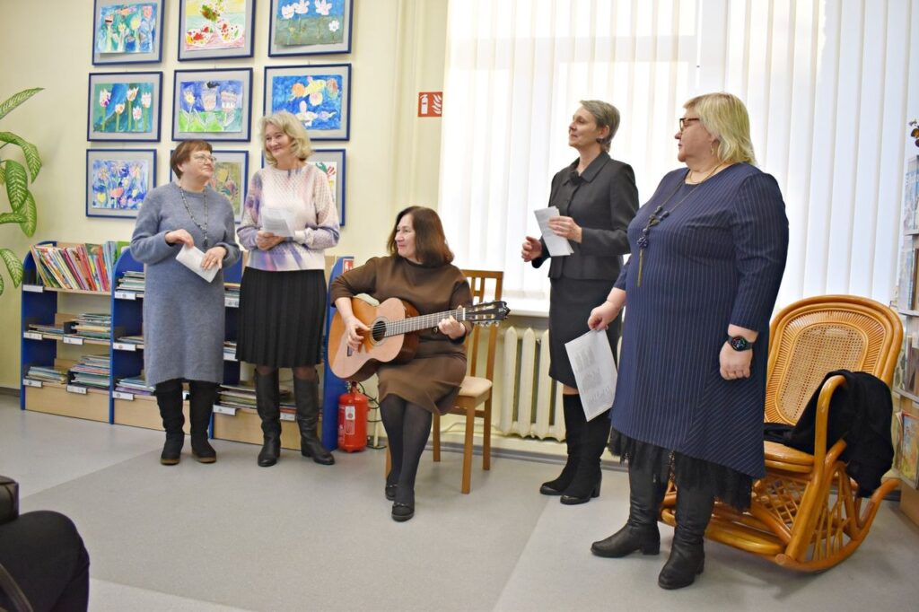 Moeris sėdėdama groja gitara, kitos moterys stovėdamos dainuoja