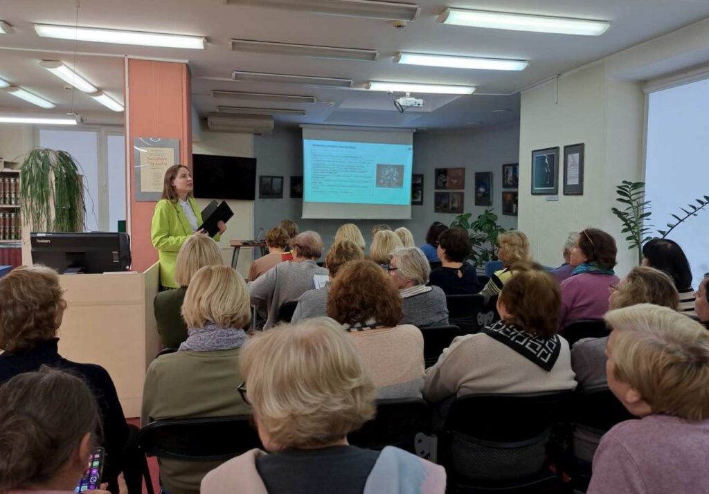 Pilna salėje sėdinčių žiūrovų klausosi lektorės